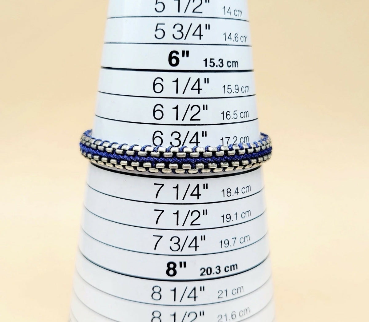 A close-up of a silver chain bracelet with blue and white striped woven bands, displayed on a white cone-shaped ring sizer with printed inch and centimeter measurements.