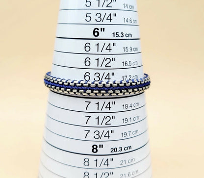 A close-up of a silver chain bracelet with blue and white striped woven bands, displayed on a white cone-shaped ring sizer with printed inch and centimeter measurements.