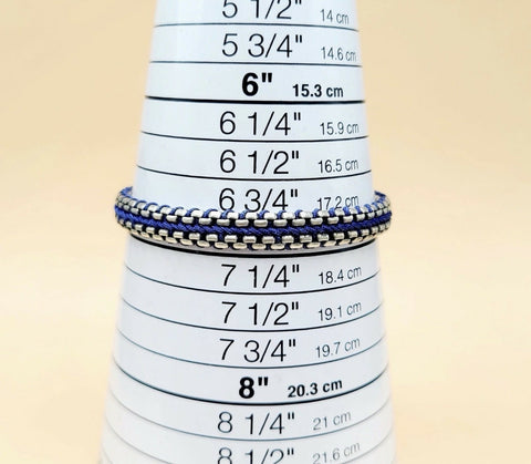 A close-up of a silver chain bracelet with blue and white striped woven bands, displayed on a white cone-shaped ring sizer with printed inch and centimeter measurements.