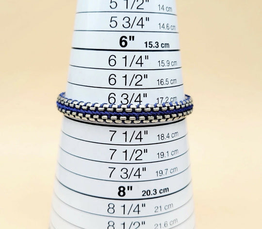 A close-up of a silver chain bracelet with blue and white striped woven bands, displayed on a white cone-shaped ring sizer with printed inch and centimeter measurements.