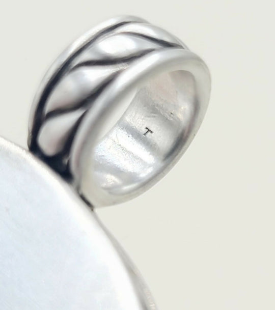 A close-up of a silver ring with a textured, wavy band design, resting on a plain light background.