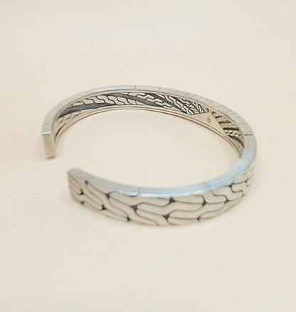 A silver-toned open cuff bracelet with a textured, wavy geometric pattern on the outer surface and engraved details on the inner band, shown against a plain light beige background.