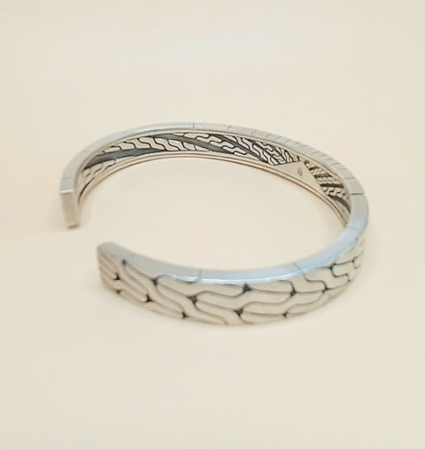 A silver-toned open cuff bracelet with a textured, wavy geometric pattern on the outer surface and engraved details on the inner band, shown against a plain light beige background.