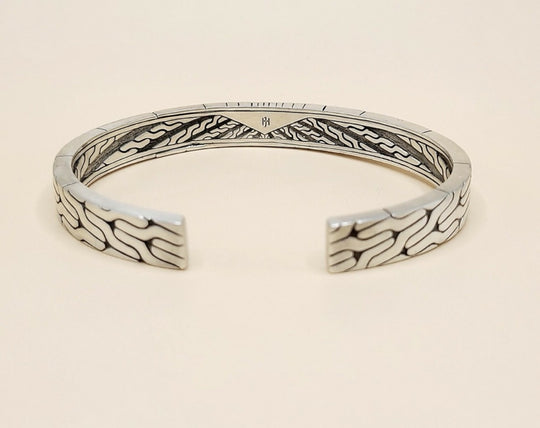 A sterling silver cuff bracelet with intricate engraved wave patterns on the exterior and interior, displayed on a light beige background.