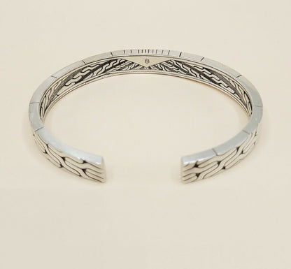 A sterling silver cuff bracelet with a textured, geometric wave pattern on the outer surface and a polished inner band, shown on a light beige background.