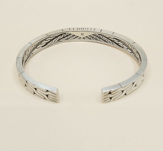 A sterling silver cuff bracelet with a textured, geometric wave pattern on the outer surface and a polished inner band, shown on a light beige background.