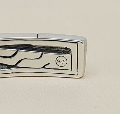 Close-up of a silver-toned metal jewelry piece, likely a cufflink or decorative clasp, with a polished rectangular face, engraved abstract lines, and a small circular '925' sterling silver hallmark on a warm beige background.