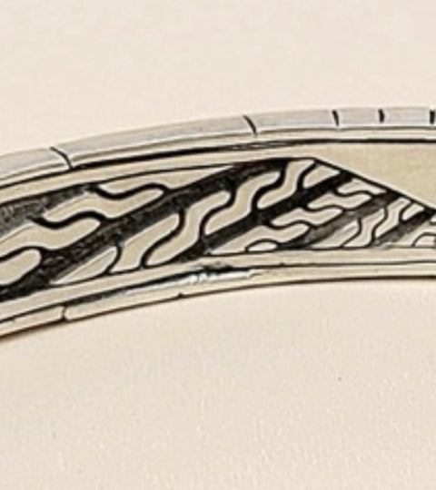 A close-up of a silver-toned metal bangle bracelet featuring a detailed black engraved pattern of wavy lines and geometric shapes.